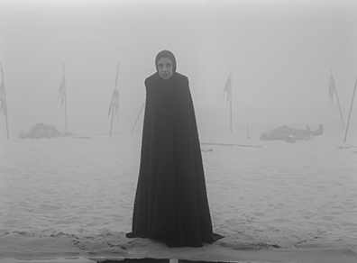 Macbeth-Witch 0105 Kathryn Hunter as a witch in black hood and ground-lengh robe on a foggy beach with in the backgroun bodies lying on the sand and six flags stuck into the ground (black and white photograph).