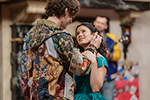 Romeo with back to us is wearing a multicolor hoodie, his right hand stroking her hair as she gaes up into his eyes, her left hand on his right arm. She's wearing a turquoise dress. A man in a blue and yellow letter jacket is playing guitar at the back of the stage.