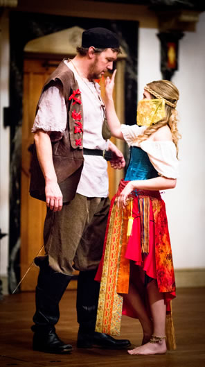 Wilmore in black hat, peasant outfit with white tunic, brown vest (featurig red ribbons) brown pants and knee-high boots, and Hellena dressed in bright blue, red, and orange gipsy dress, yellow veil around her face, and a finger pointed at Willmore's nose.