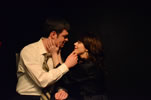 Mosby in light yellow oxford shirt and gold striped tie sitting in a chair strokes the chin of Alice, with auburn hair and in a black robe, kneeling on the floor as she caresses his neck