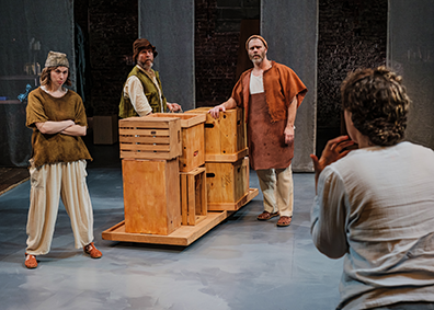 FIA-2-Fishermen Pericles, with his pack to us at the right, pleads with the three fishermen in rustic wear and standing next to a platform of stacked crates.