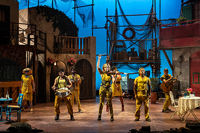 STC-Comedy_Errors-Musicians-089 The musicians in various yellow garb stand lined up across the width of the stage from cafe table and chair on the left to a home's tableclothed table and chair to the right. Behind them is the set of Ephesus, with building facades featuring doors and balconys, and a scaffold at the back covered in fishing nets and crates with a boat under construction underneath. The back screen is a twilight blue.