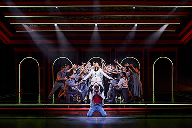 STC Evita-Ensemble Eva Peron in a black trimmed white business jacket and dress stands with arms outstretched in a spotlight as the gray-clothed people of Argentina surround her in supplication while Che in red shirt and denim jacket and jeans knees with prayerful hands upraised at the front of the stage
