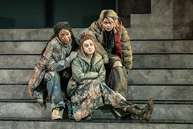 STC-Macbeth-Witches-0171 The three witches huddle on the steps to the bunker watching action toward the camera. They are wearing a colorful collection of baggy clothes and oversize coats,