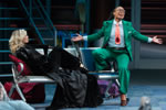 Viola, wearing a green jacket and pants, blue shirt, and bright orange tie with a giant ladybug at the bottom, sits on the arm at one end of a three-seat airport lobby bench, one foot on the ground, one on a seat, arms outspread as she talks to the air while Olivia, stretched out on the other end of the bench and wearing black pant suit and floor-length black coat watches in fascination.